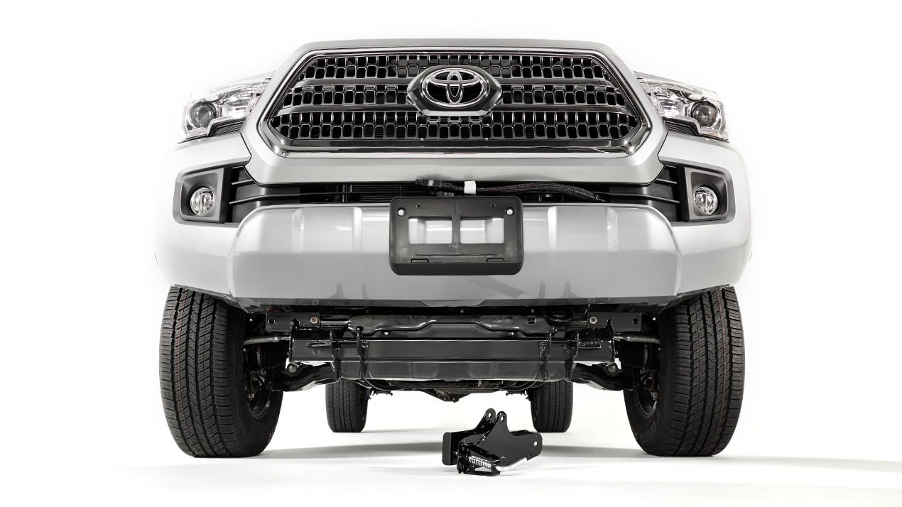 Front view of a white Toyota truck with an empty license plate holder, shown against a white background. A detached metal vehicle part is placed on the floor beneath the front bumper.
