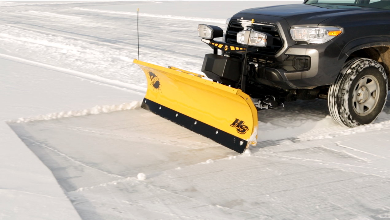 A pickup truck with a mounted yellow snow plow is clearing snow from a road, pushing it to the side and revealing the pavement underneath.
