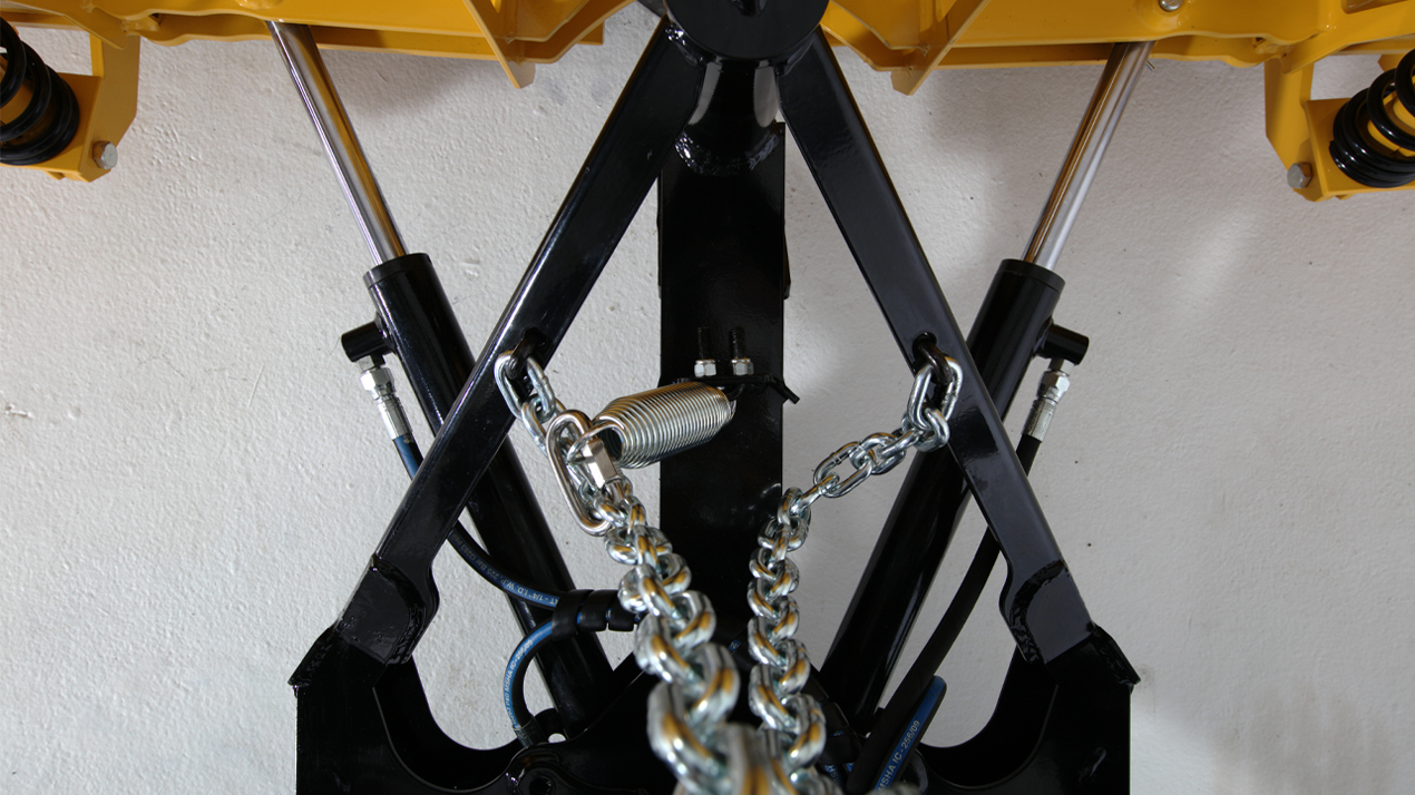 Close-up view of hydraulic machinery components featuring black metal arms, silver hydraulic cylinders, coiled spring, and a thick metal chain, all attached to a yellow structure against a white background.