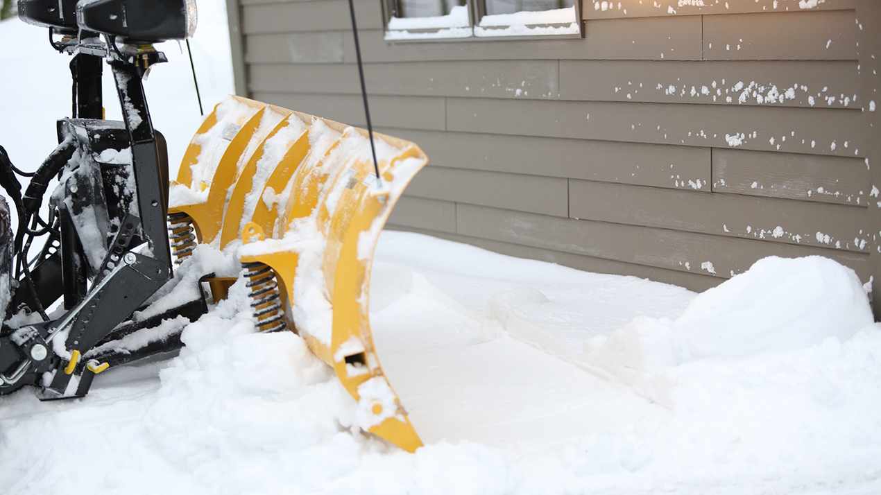 A yellow snow plow clears fresh snow near the side of a house with beige siding and snow-covered windows. Snow is piled up along the building and around the plow.