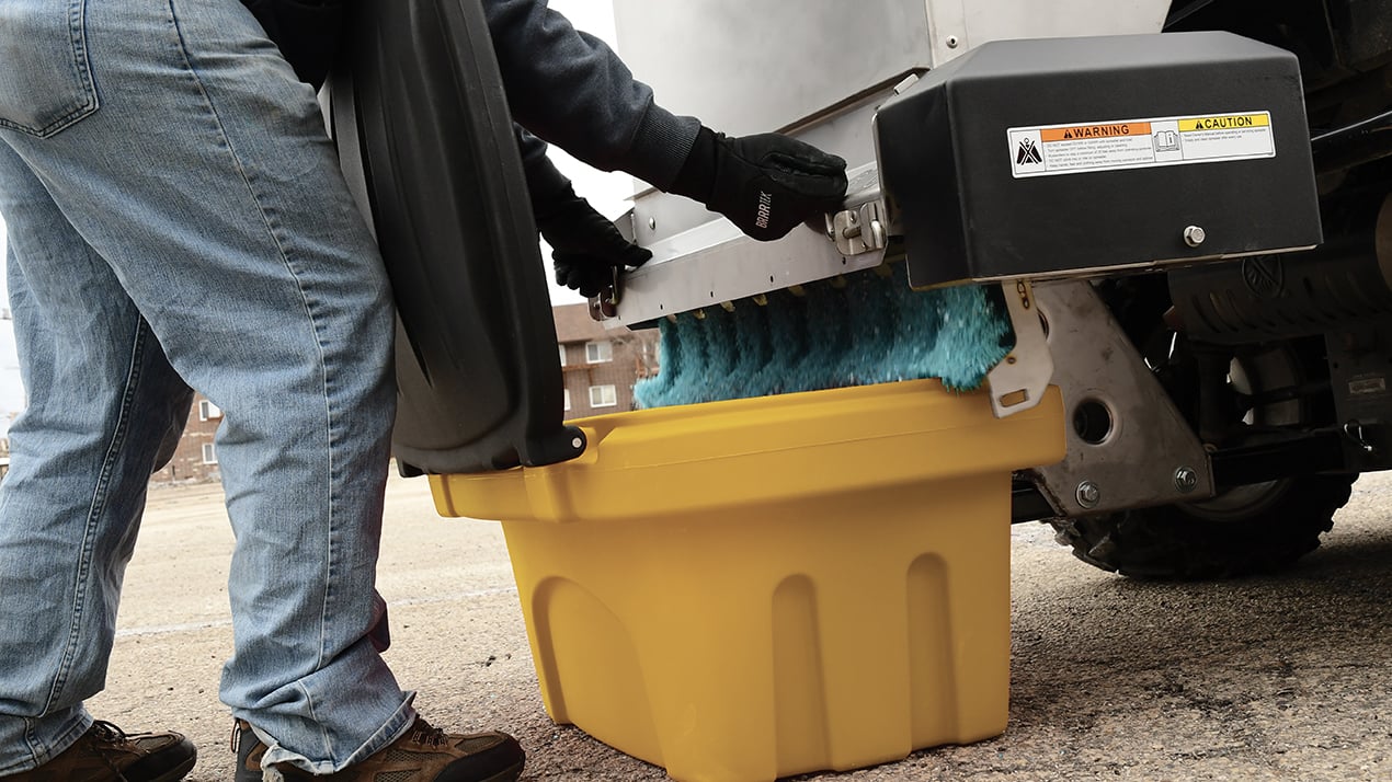 A person in jeans and gloves empties blue bristles from a street sweeper into a large yellow bin on a paved surface.