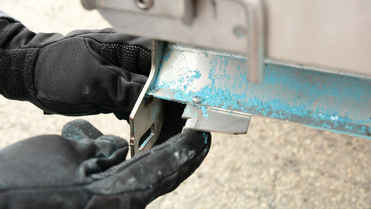 A person wearing black gloves is handling a metal part with chipped blue paint, possibly inspecting or repairing a piece of equipment outdoors.