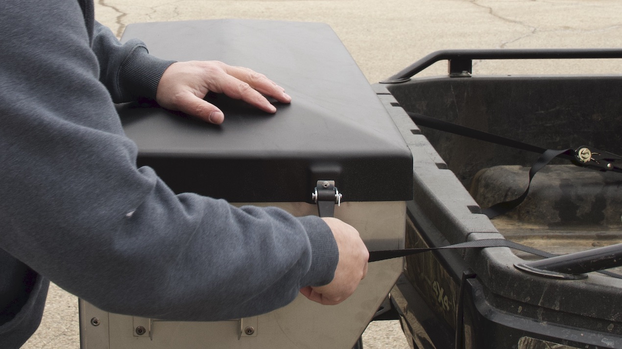 A person in a gray sweatshirt secures a black storage box to the back of a utility vehicle using a strap.