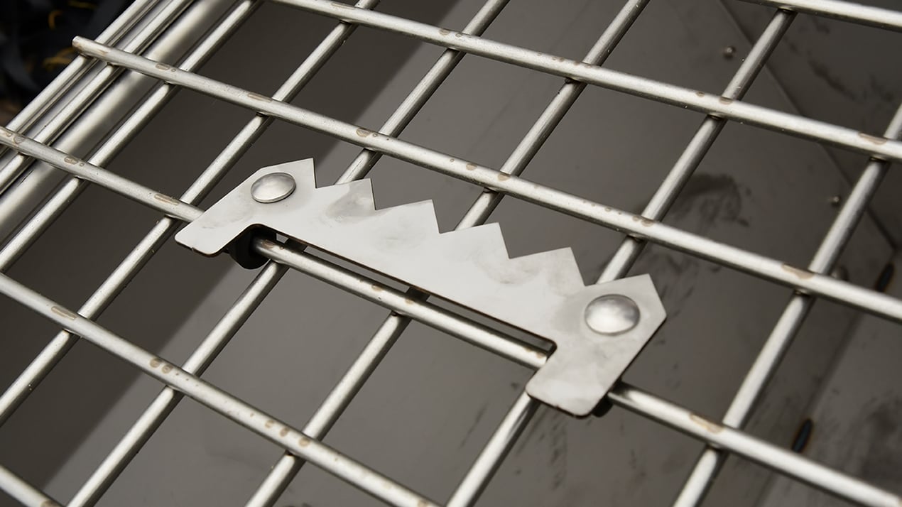 Close-up of a metal locking mechanism with jagged teeth on a metal wire cage or crate. The background shows the inside of the enclosure.