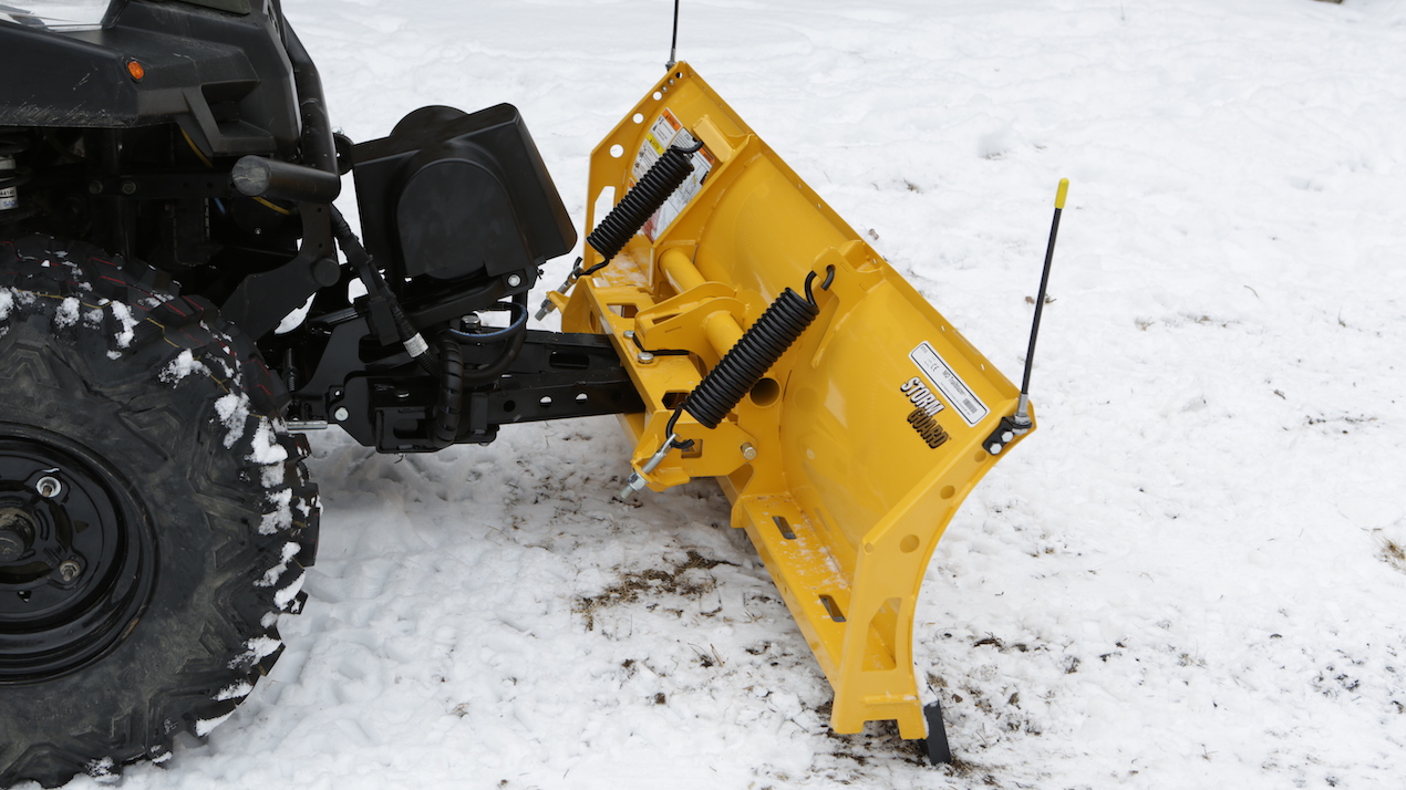 A close-up of a yellow snowplow blade attached to the front of a black vehicle, positioned on a snowy ground. The snowplow features coiled springs and metal mounting hardware.
