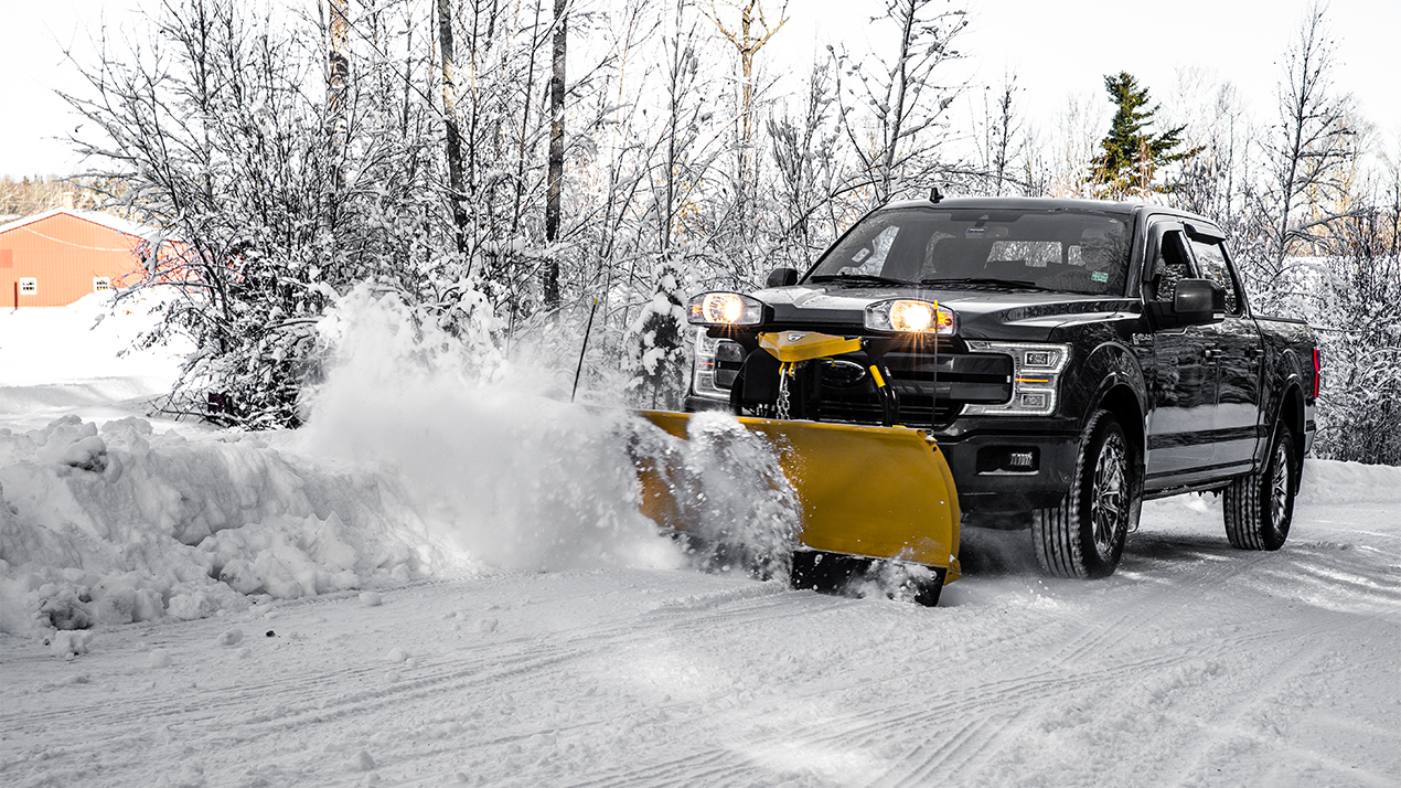 A black pickup truck equipped with a yellow snow plow clears snow from a road, surrounded by snowy trees and a building in the background during winter.