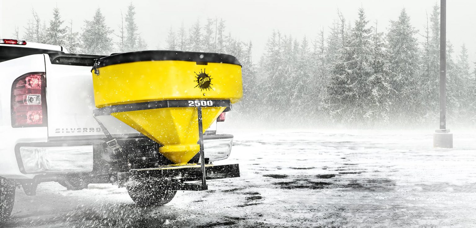 A yellow salt spreader attached to the back of a white pickup truck disperses salt on a snowy, icy road. Snow is falling, and pine trees are visible in the foggy background.