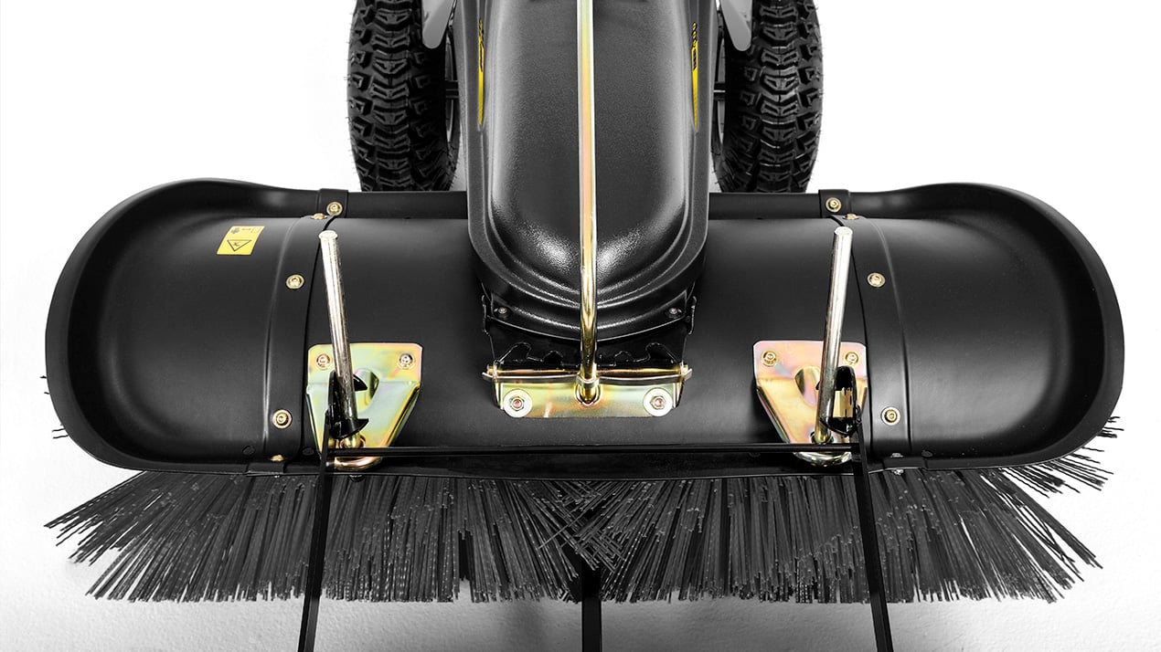 Close-up view of a street sweeper attachment with stiff black bristles and metal components, mounted on a vehicle with large, treaded tires in the background.