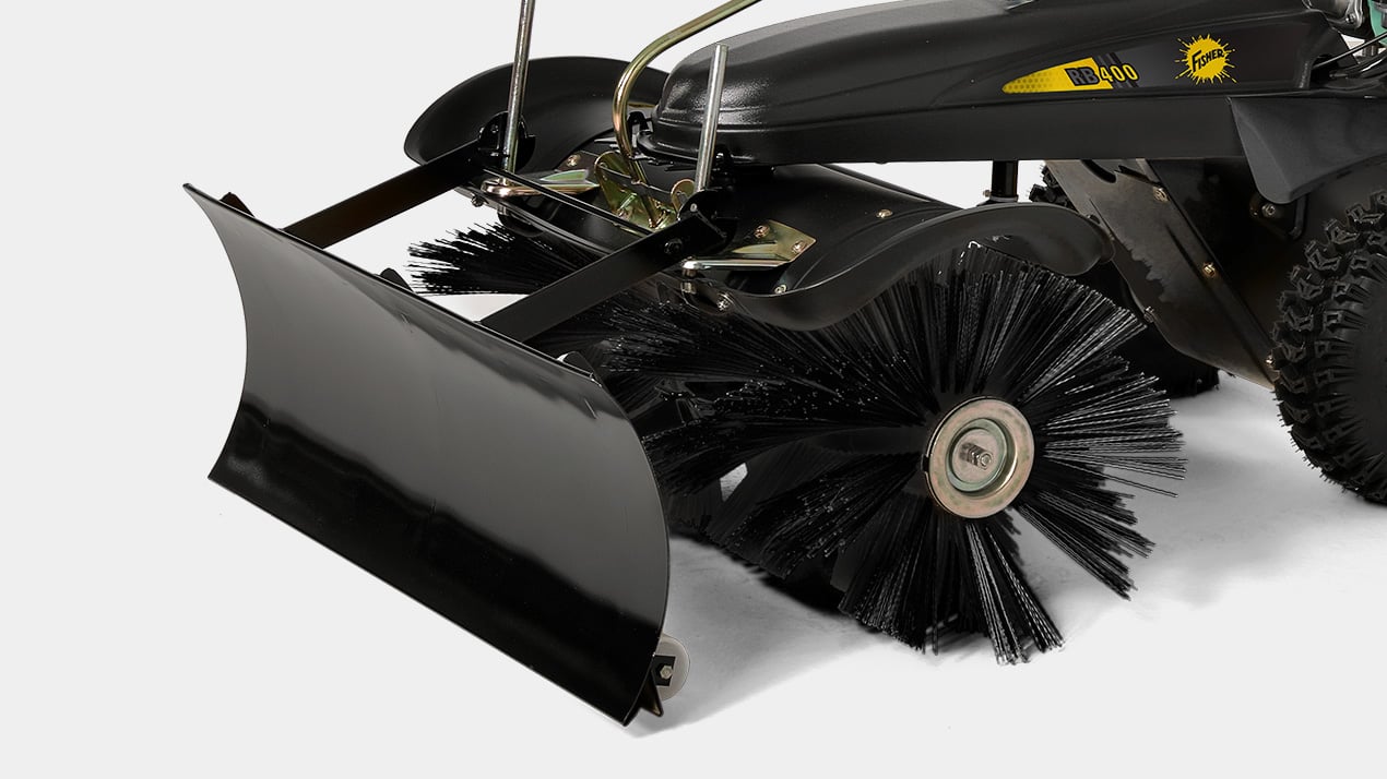 A close-up view of a mechanical outdoor sweeper with a large rotating brush and an attached black snow plow blade on the front.