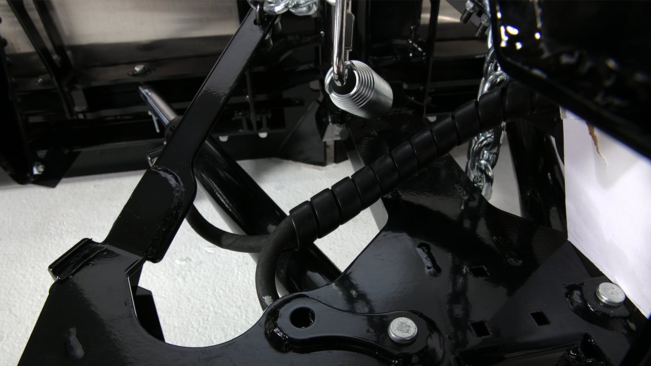Close-up of black metal machinery parts with bolts, a coiled cable, and hydraulic components, set against a white background.