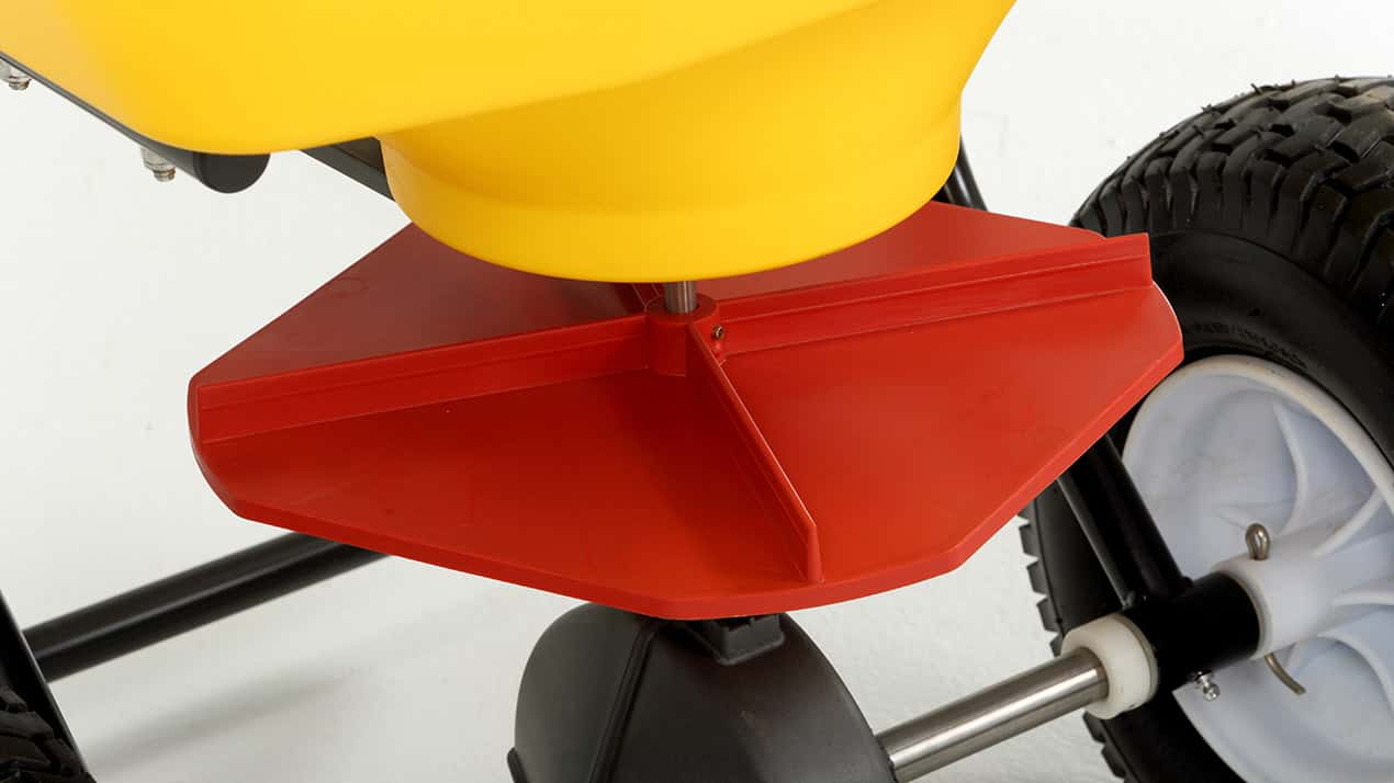 Close-up view of a red plastic spinning disc attached below a yellow container, with part of a black metal frame and large rubber wheels visible, likely part of a garden seed or fertilizer spreader.