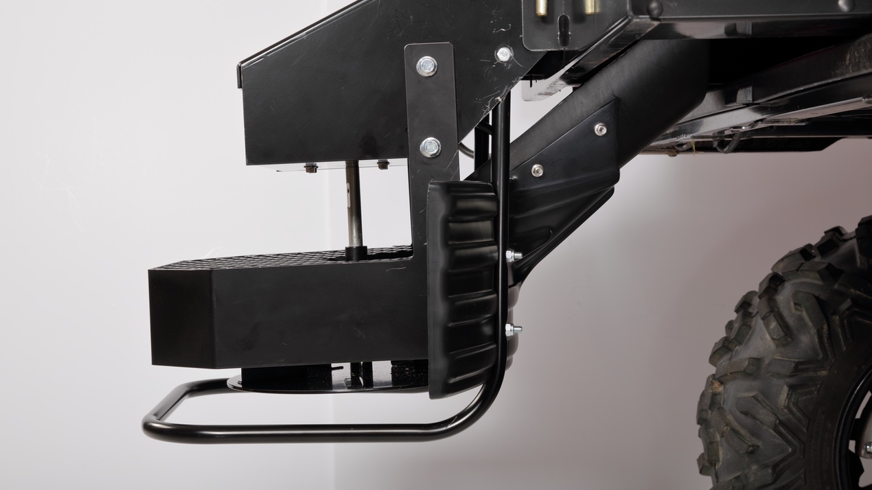 Close-up view of a heavy-duty metal vehicle step attached to the frame of an off-road truck, showing black metal components, bolts, and part of a large rugged tire on the right.