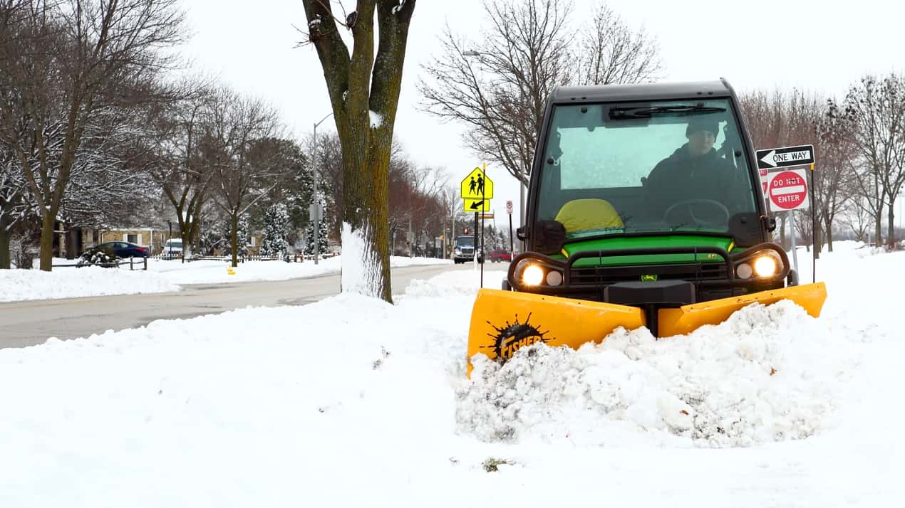 A utility vehicle with a yellow snow plow attachment clears snow from a sidewalk in a residential neighborhood on a winter day. Snow covers the ground, and trees line the street.
