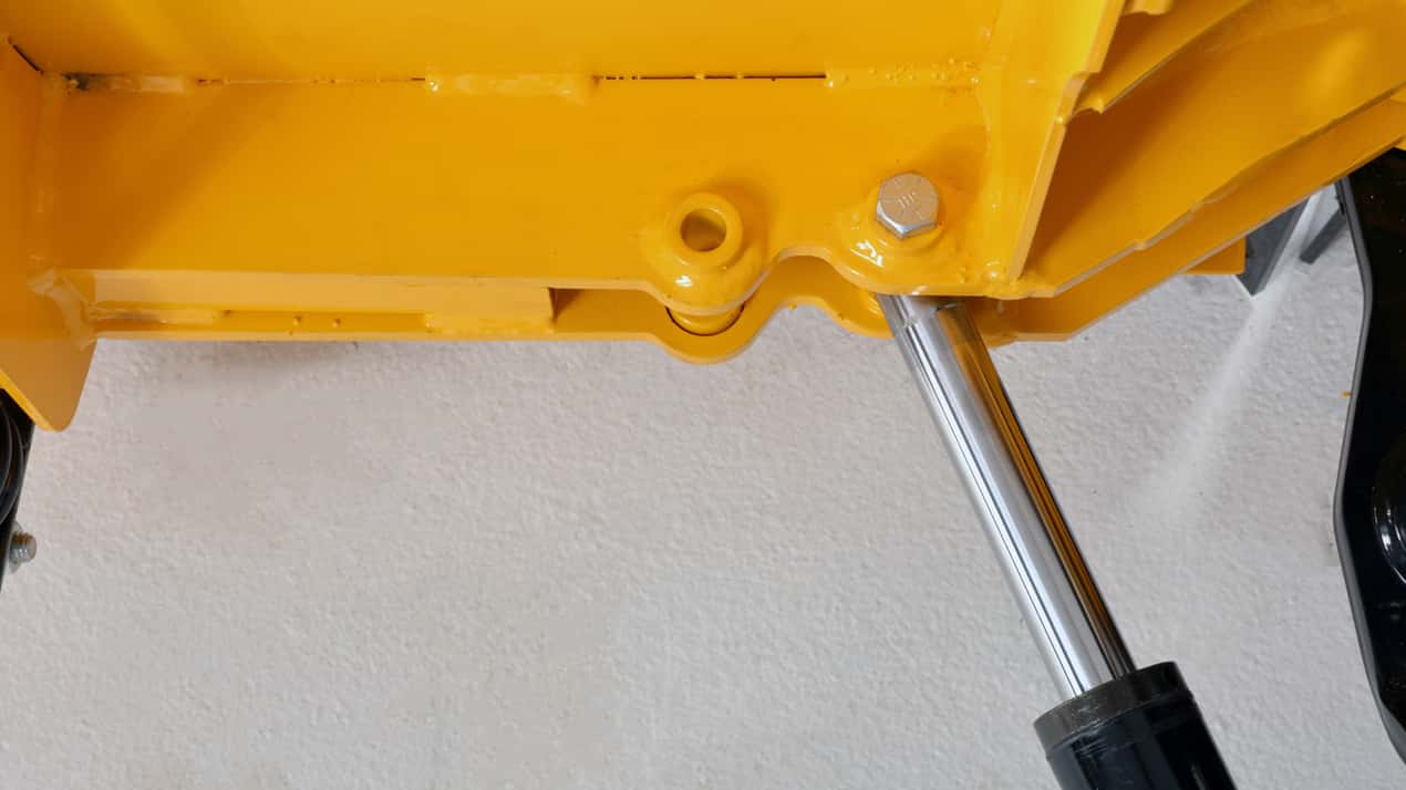 Close-up of a yellow metal structure with a bolt and a shiny hydraulic piston or cylinder attached, set against a white background.