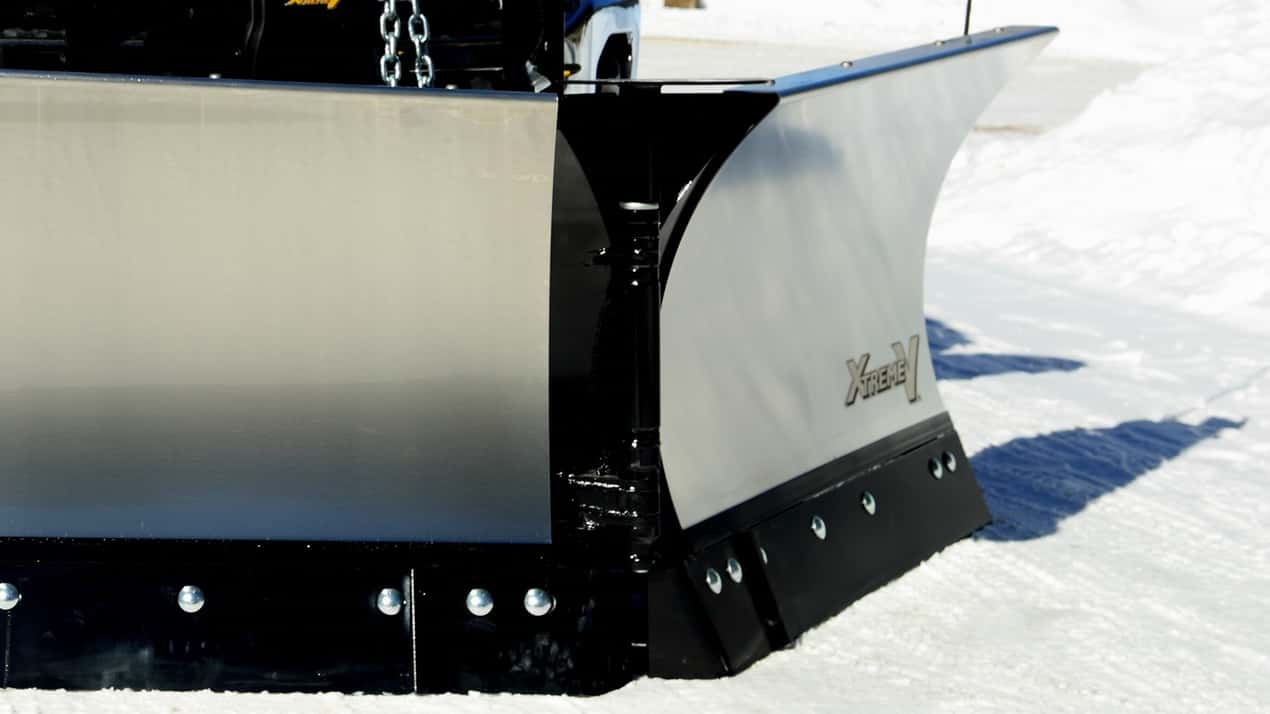 Close-up of a large snow plow attached to the front of a vehicle, clearing snow on a bright winter day. The plow is metallic with black edges and bolts along the bottom.
