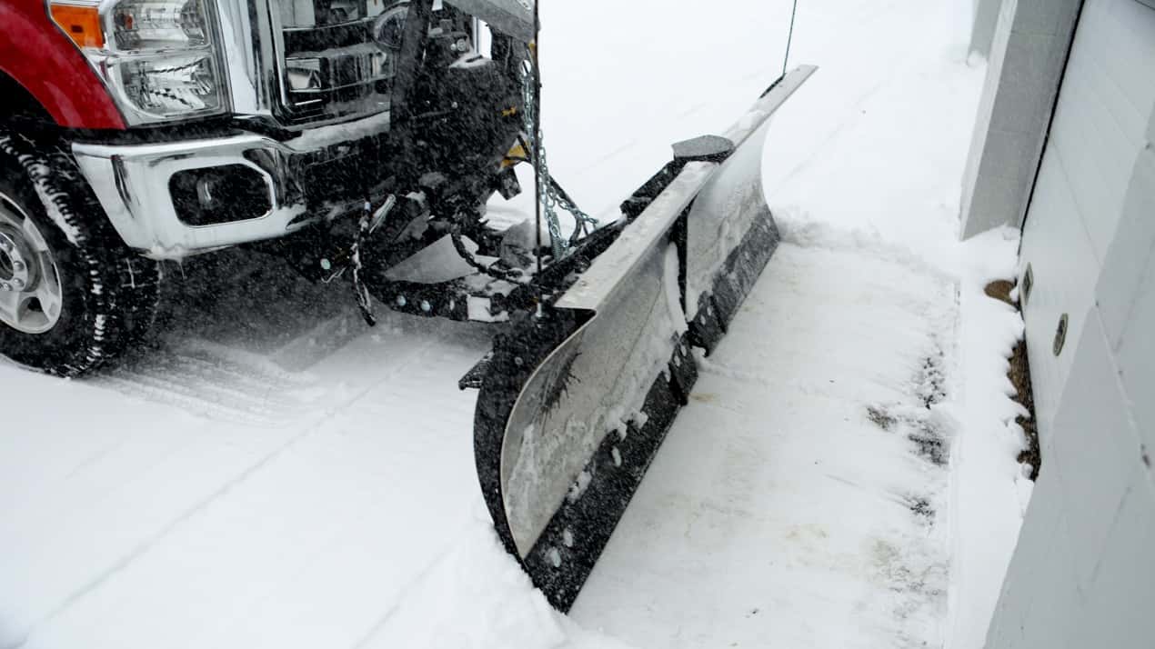 A red truck with a large snow plow clears fresh snow from a driveway next to a white building during winter.