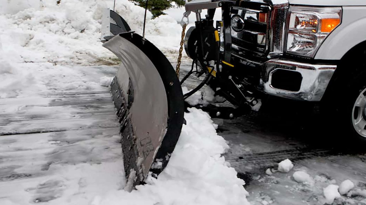 A truck with a plow attached is clearing snow from a paved surface, pushing aside a pile of fresh snow. The scene is snowy and wintry, with the truck mostly visible from the front side.