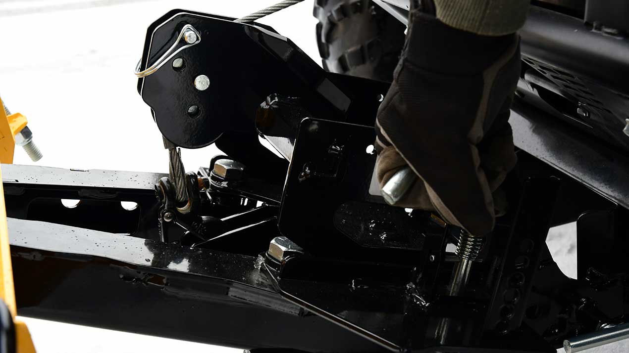 A person wearing gloves adjusts a steel cable and mechanical winch assembly attached to a black metal frame, likely part of heavy machinery or a vehicle, on a snowy background.