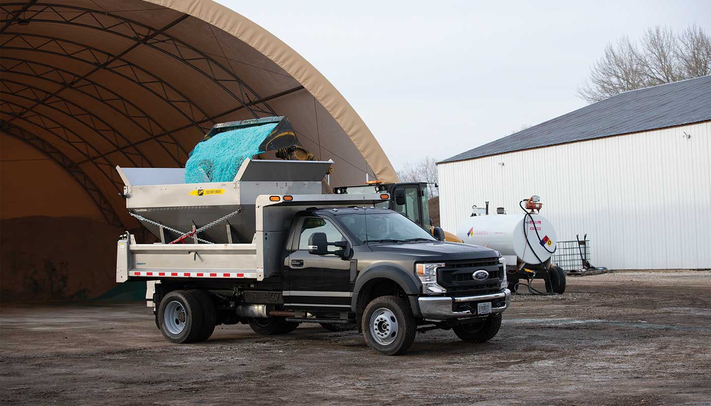 A black dump truck is parked outside a large storage tent, with green salt or de-icing material being loaded into its rear spreader from a front loader. A white utility tank is visible in the background.