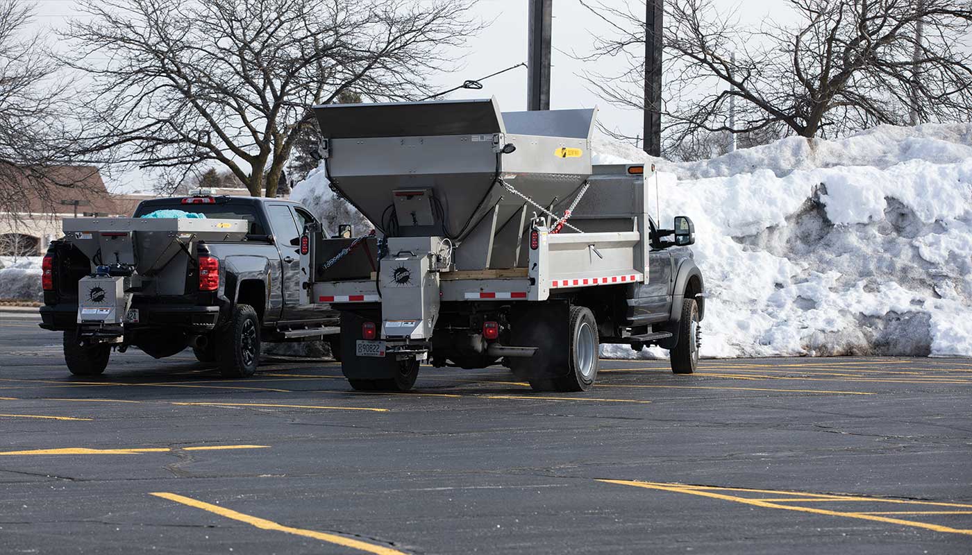 Two pickup trucks with salt spreaders attached are parked in a lot with large piles of snow in the background. The sky is overcast and bare trees line the lot. Yellow parking lines are visible on the asphalt.