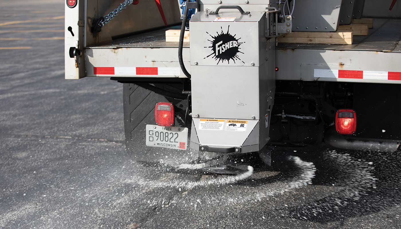 A truck with a Fisher salt spreader mounted on the back disperses salt onto an asphalt surface, preparing the area for icy conditions. The truck’s rear lights are on and salt is visibly spreading below.