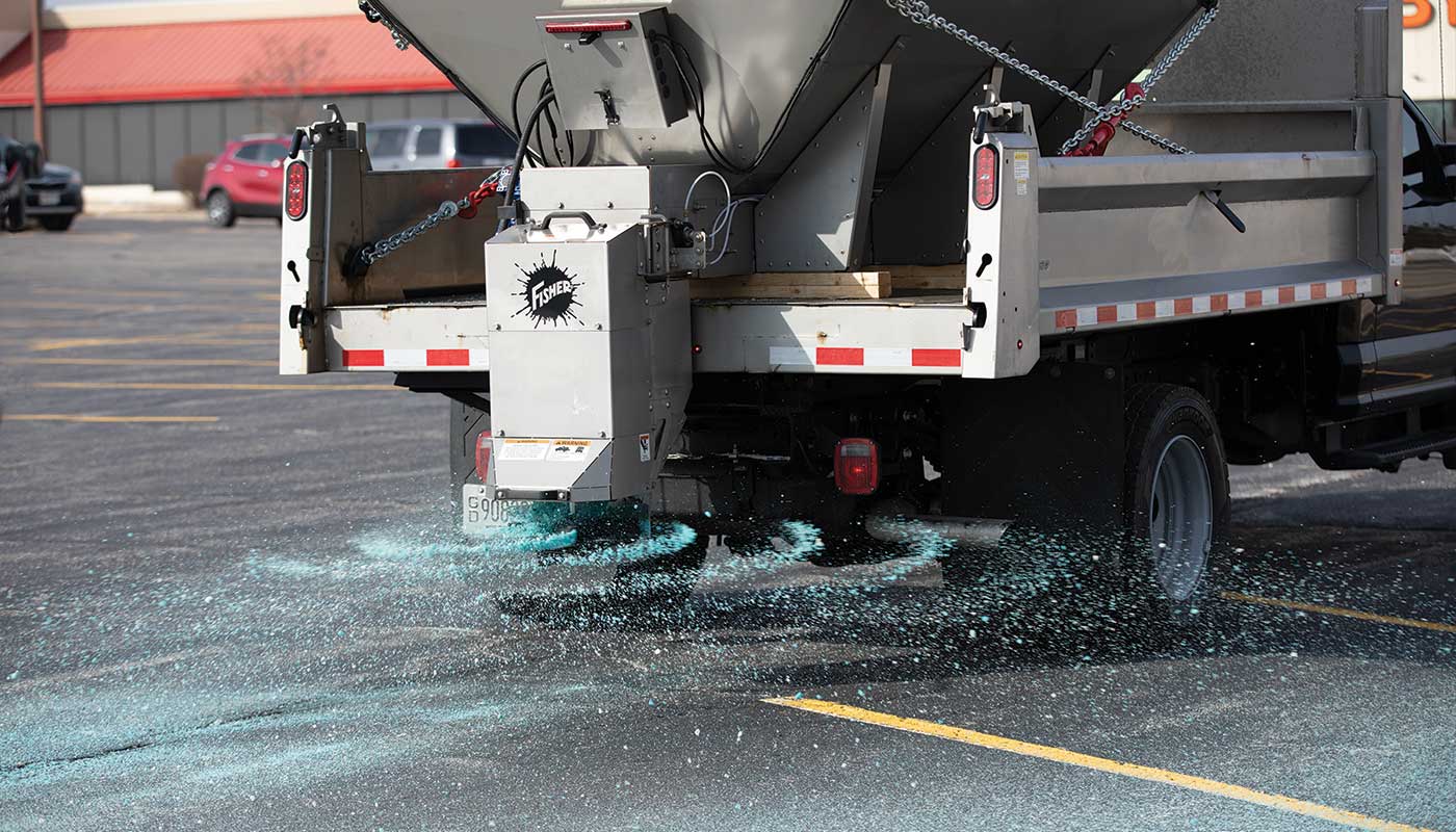 A truck spreads blue de-icing salt on a parking lot, with salt visibly being dispensed from the rear of the vehicle and scattering across the asphalt.