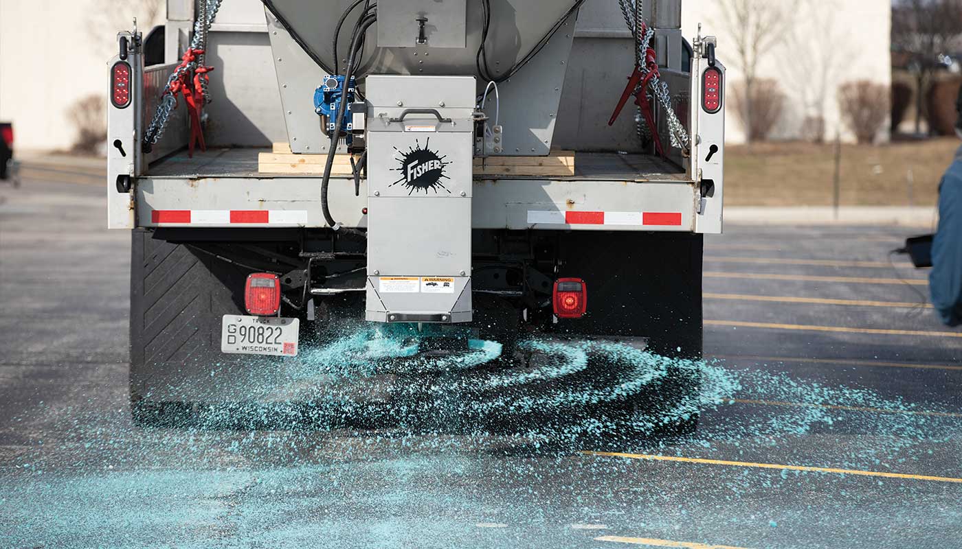 A truck spreads blue de-icing salt on an asphalt parking lot, with swirling patterns forming behind the vehicle. The scene appears to be in winter, with bare trees and no visible snow.