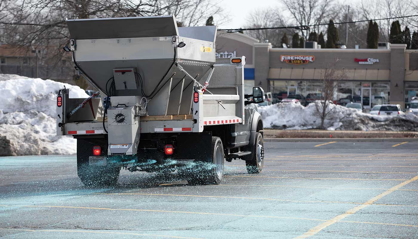 A truck spreads blue road salt in a snowy parking lot in front of shops, including Firehouse Subs, on a winter day with snow piles in the background.