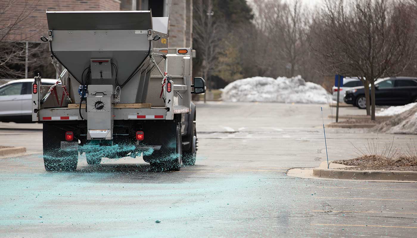 A truck spreads blue de-icing salt onto a parking lot, with piles of snow and bare trees visible in the background on a cloudy day.