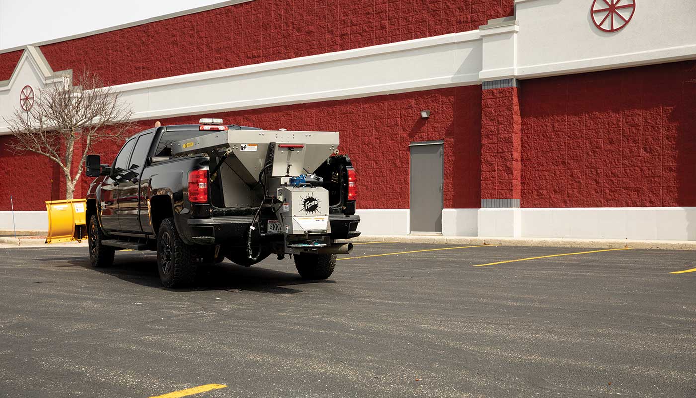 A black pickup truck with equipment mounted in its bed is parked in an empty lot near a large red and white building with decorative accents. A yellow container sits by a leafless tree in the corner.