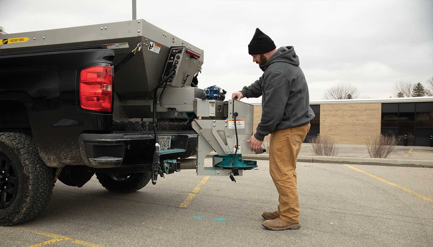 A man in a black beanie and gray hoodie operates a salt spreader attached to the back of a black pickup truck in a parking lot on a cloudy day.