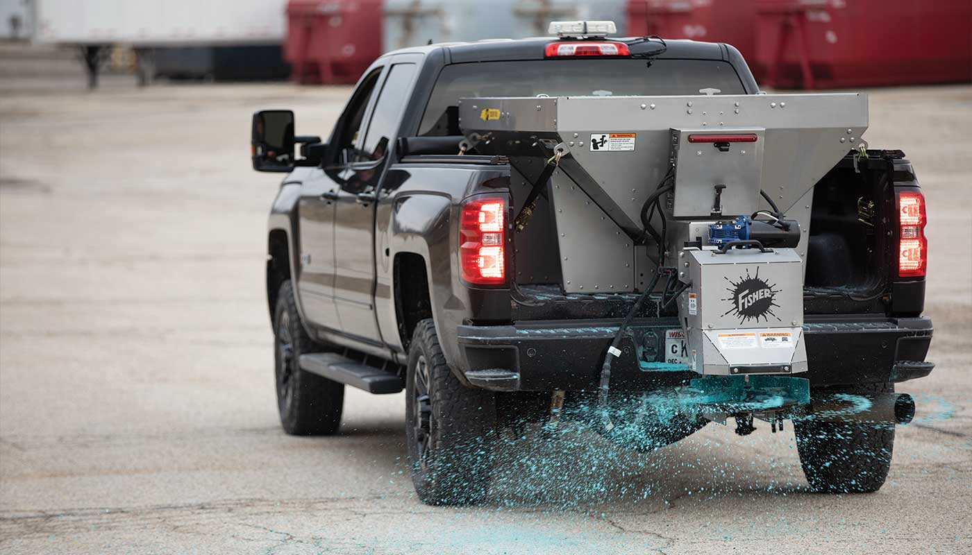 A black pickup truck is spreading blue salt or de-icing material onto the ground from a mounted spreader attached to its rear, in an industrial or parking lot setting.
