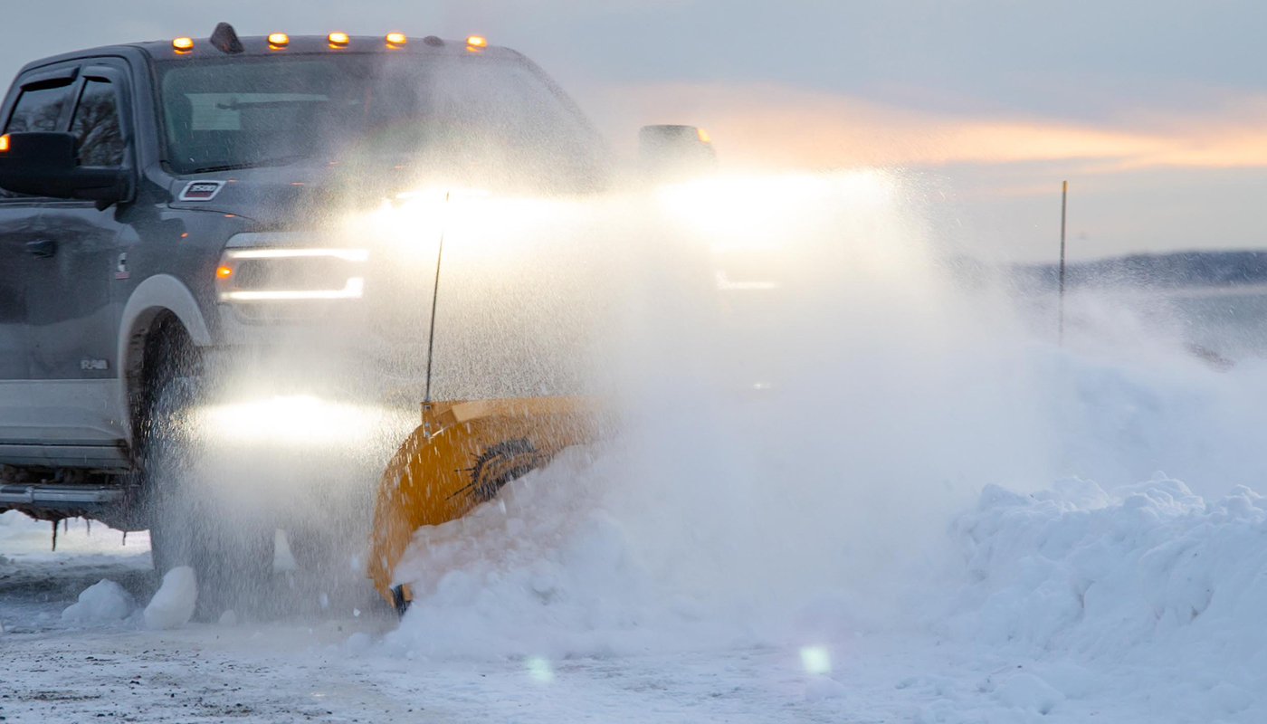 A pickup truck with headlights on is plowing snow at sunset, pushing a large pile of snow with a bright yellow plow blade, while snow blows up around the vehicle.