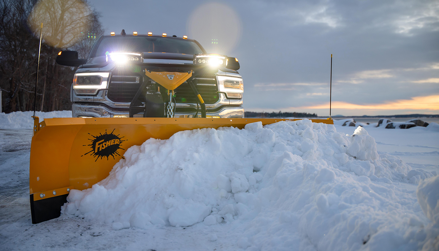 A pickup truck with headlights on uses a large yellow Fisher snow plow to clear a pile of snow from a road or driveway at sunrise or sunset in a snowy landscape.