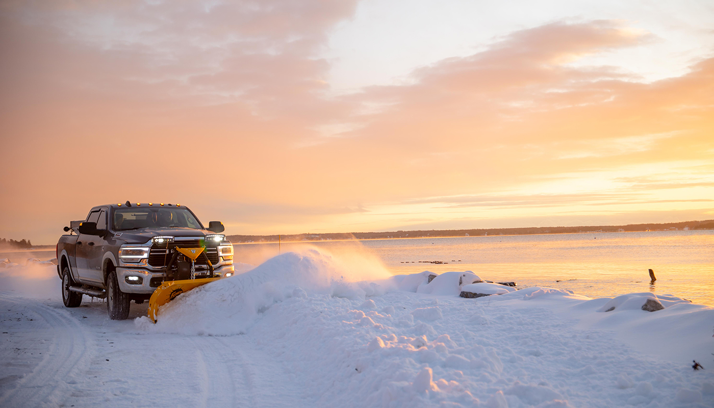 A pickup truck with a yellow snow plow attachment clears snow on a scenic, snowy road near a large frozen lake at sunrise, with warm orange and pink tones illuminating the sky.
