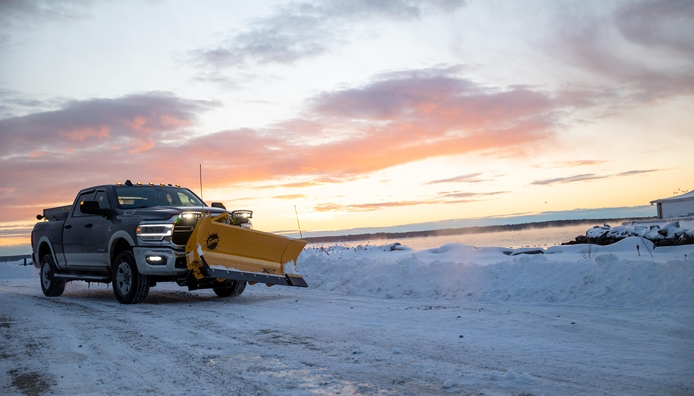 A black pickup truck with a yellow snowplow attachment clears a snow-covered road at sunrise, with colorful clouds in the sky and snowy landscape in the background.