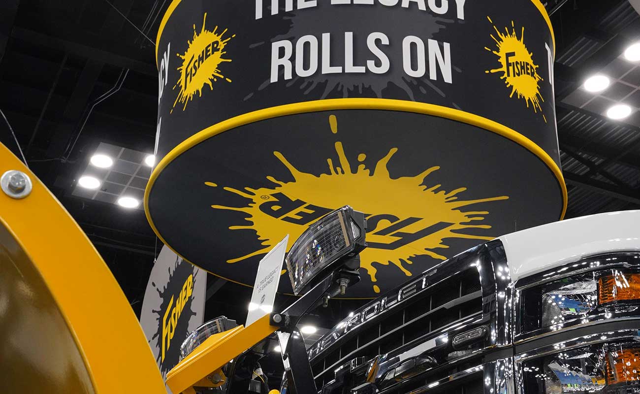 Close-up of a yellow and black Fisher snowplow display at work truck week, with a large circular sign above reading THE LEGACY ROLLS ON and bold yellow splatter graphics in the background.